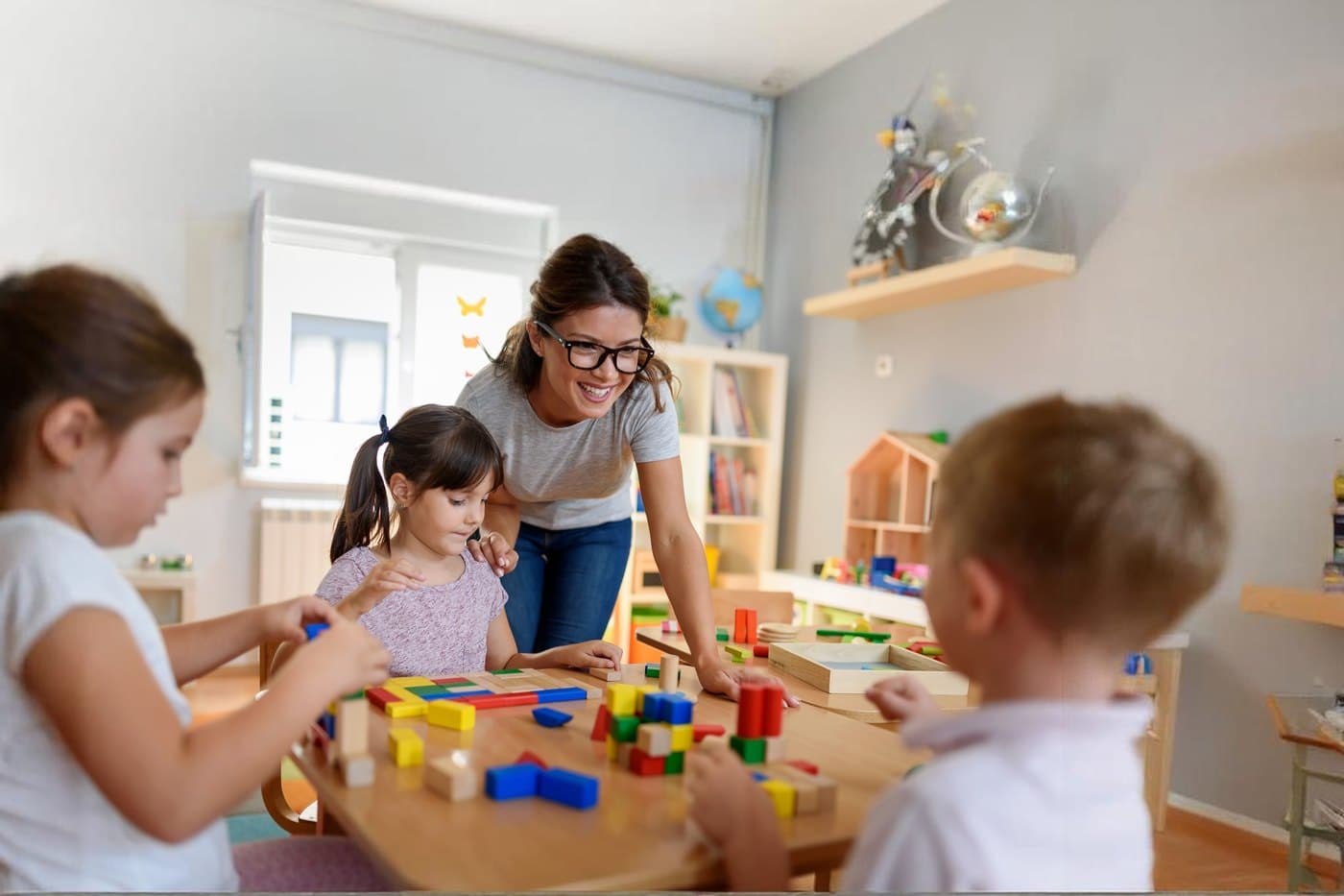 A smiling teacher building with wooden blocks alongside three children at a classroom table